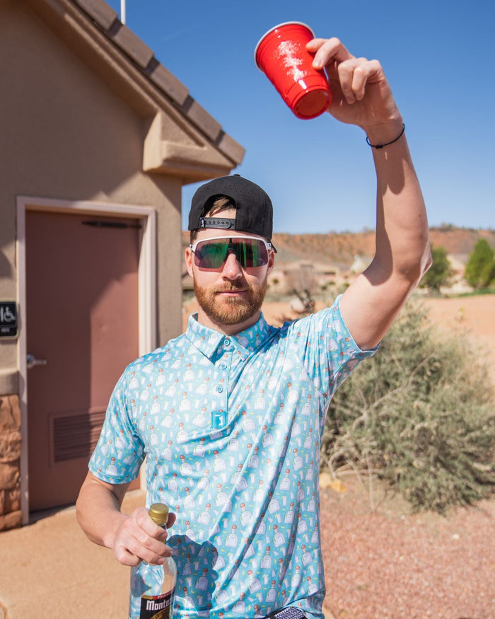 Wearing the Tequila - Polo in an athletic fit, a bearded man with a backward black cap and reflective sunglasses stands outdoors by a desert-view building, raising a red cup and holding a bottle.