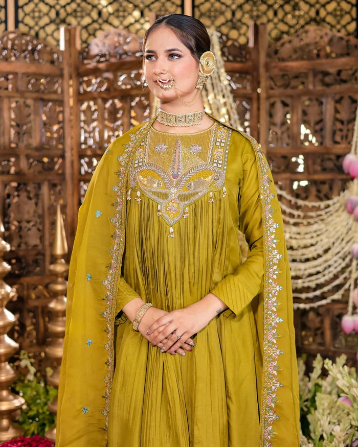Woman in a green traditional outfit with intricate patterns, standing against a decorative background.
