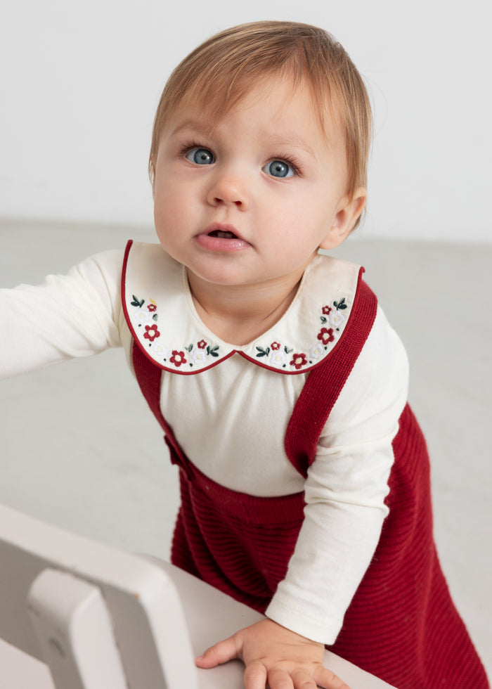 Baby wearing white bodystocking with floral embroidered collar and Christmas red suspender dress