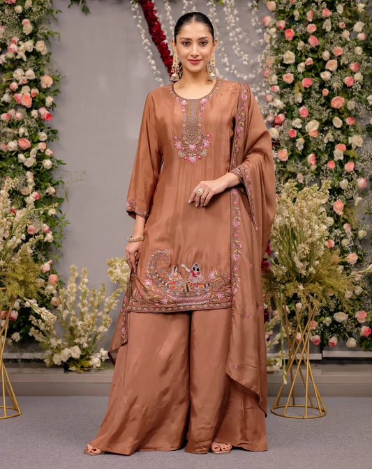 Woman in a brown embroidered traditional outfit standing in front of floral decorations.