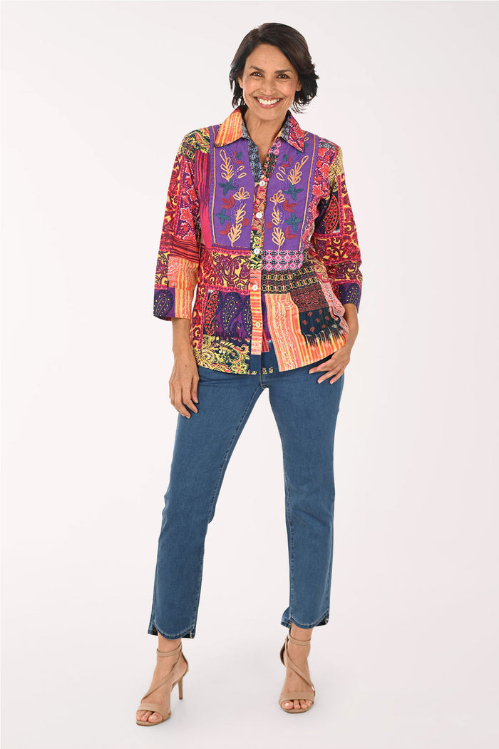 A woman modeling a vibrant, multicolored patchwork button-up shirt with blue jeans and beige high-heeled sandals, standing against a plain white studio background.