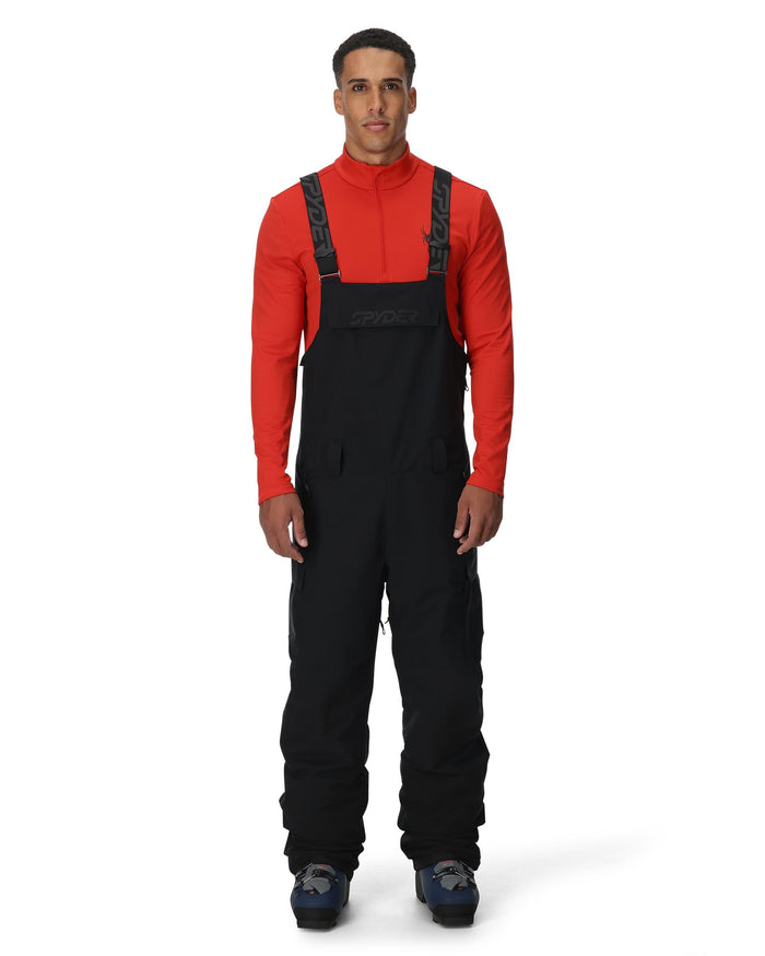 Man wearing a red long-sleeve shirt and black bib overalls on a white background