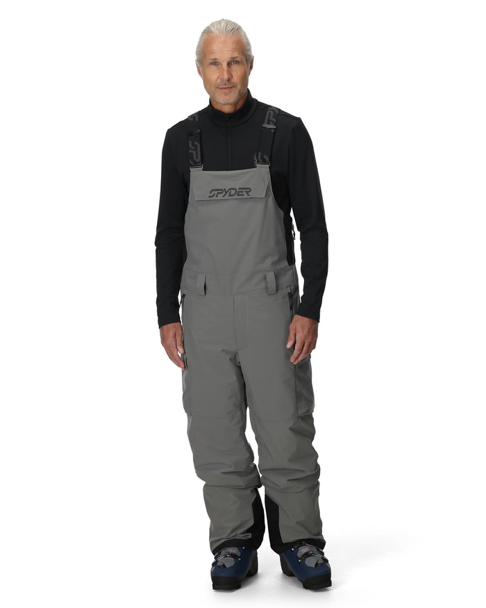 Man wearing gray bib overalls with Spyder logo on a white background
