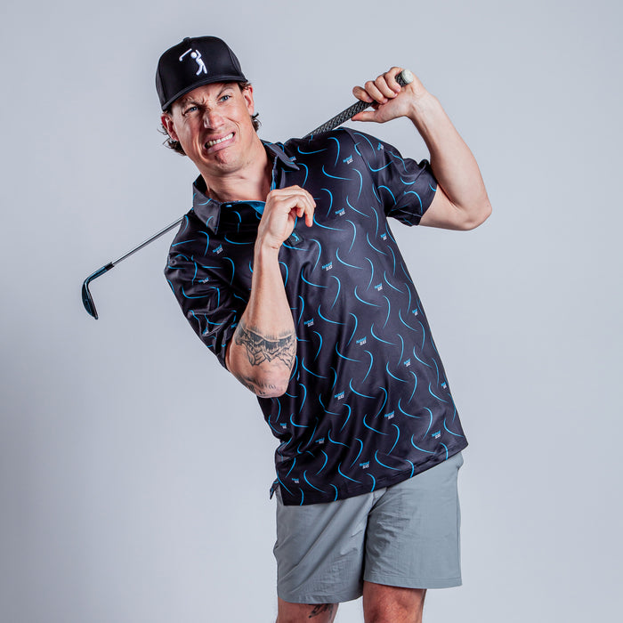 A man in a black hat wears the Natural Slice - Polo, an athletic-fit, patterned golf shirt, and gray shorts. He makes a humorous face while holding a golf club over his shoulders, showing off a tattoo. The background is plain light gray.