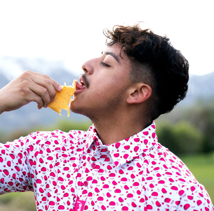 A young man with short dark hair and a trimmed beard is outside on a bright day, eating a cheesy taco while wearing the Pink Taco - Polo, a moisture-wicking white shirt featuring a pink pattern. Greenery and distant mountains blur in the background.
