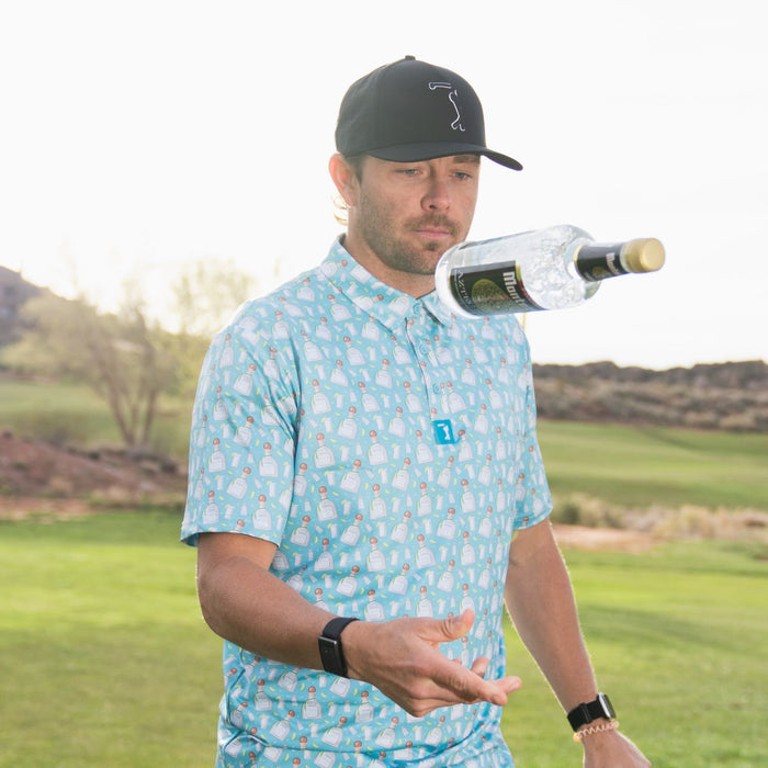 On a sunny golf course, a man in the Tequila - Polo—an athletic fit, moisture-wicking blue shirt—tosses a clear bottle in the air. He pairs it with a black cap, smartwatch, and wristband amid grass, trees, and distant hills.
