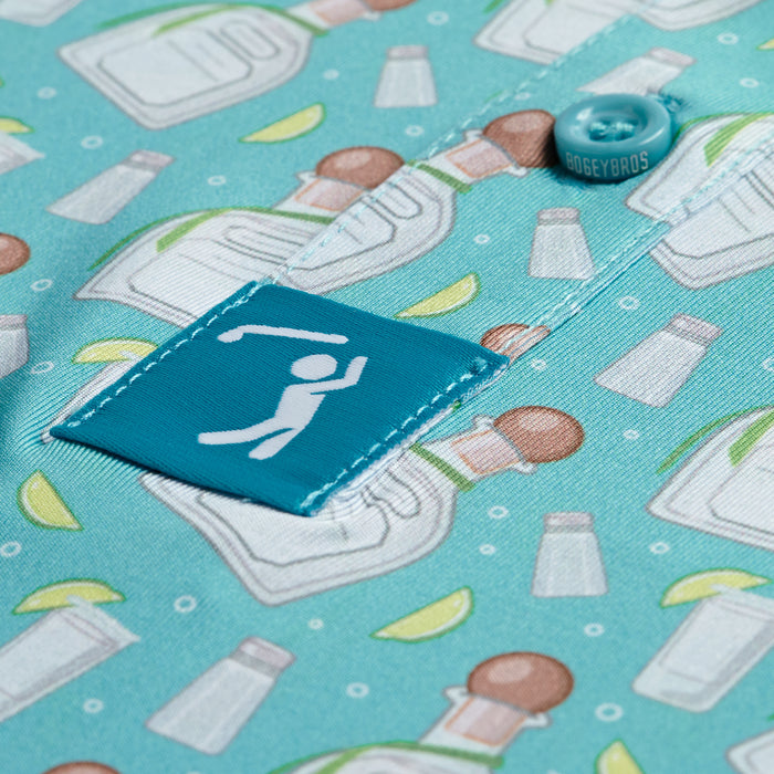 A close-up of the Tequila - Polo, an athletic fit turquoise shirt with a pattern of tequila bottles, salt shakers, and lime wedges. Features a “BOGEYBROS” button and a blue tag with a golfer silhouette.