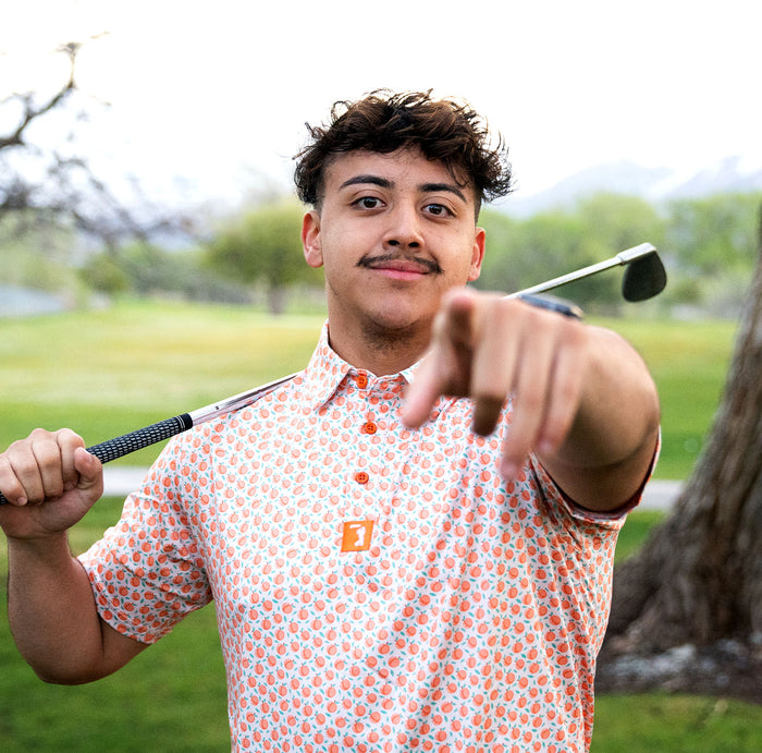 A young man with short wavy hair and a mustache smiles outdoors on a golf course, holding a club over his shoulder and pointing at the camera. He wears the Tooty Frooty - Polo, an athletic-fit shirt with UPF 50 protection.
