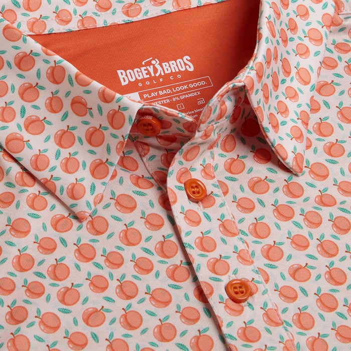 Close-up of the Tooty Frooty - Polo, an athletic-fit collared shirt with a playful all-over peach print, orange buttons, and inner collar. Perfect for summer golf days. Label: Bogey Bros Golf with washing instructions.