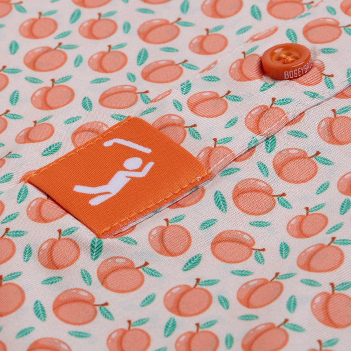 Close-up of the Tooty Frooty - Polo, showcasing a fabric patterned with small orange peaches and green leaves. The athletic fit features an orange tag with a white figure raising arms, plus an orange button engraved with BOGEY.
