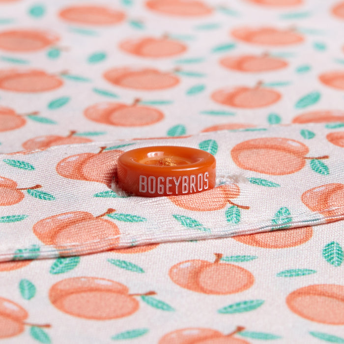 Close-up of the Tooty Frooty - Polo fabric with an orange peach and green leaf pattern, accented by an orange BOGEYBROS button on a seam. The fabric is moisture-wicking for comfort during activity.
